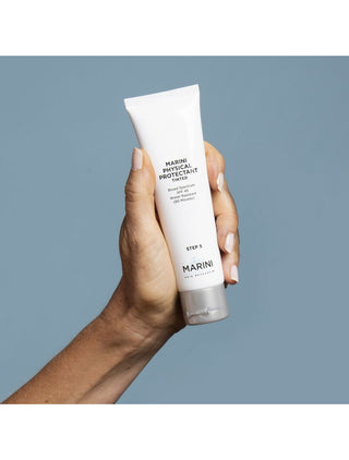Blue background with hand holding up Jan Marini Physical Protectant Tinted SPF. The perfect sunscreen to help protect skin from free radicals