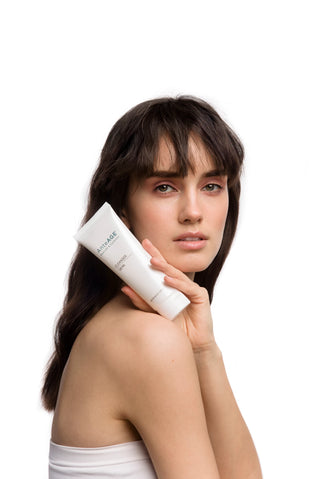 White background with a girl who has medium length dark hair and bangs looking over her shoulder and holding a bottle of the Anteage Cleanser (Skin Cleanser)