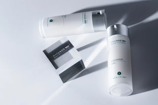A grey background with two Anteage MD bottles laying down. Each bottle is a white or clear frosted color with a grey dispenser that casts a large shadow. This product is a two part system used as a go to topical treatment after any facial procedure. These help with the healing process and improve skin health while helping skin appearance.